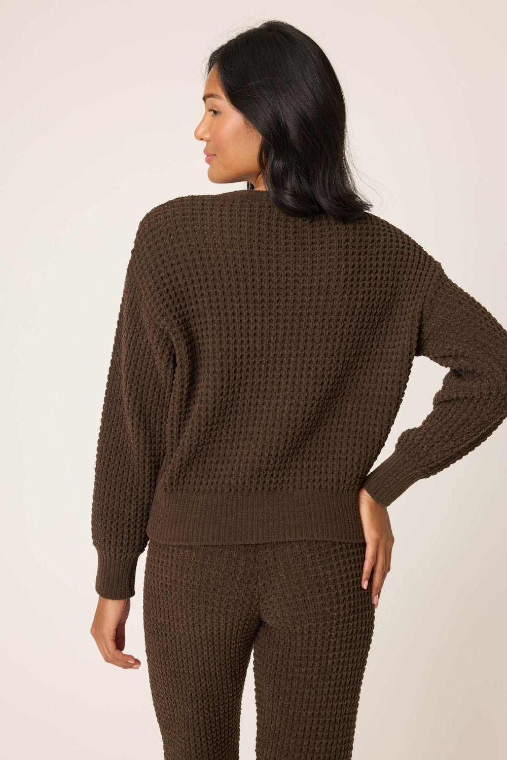 A woman faces away from the camera, wearing the P.J. Salvage Chenille Waffle Solid Chocolate Sweater with matching pants. Her right hand is on her hip, and she has medium-length dark hair. The background is plain and light-colored.