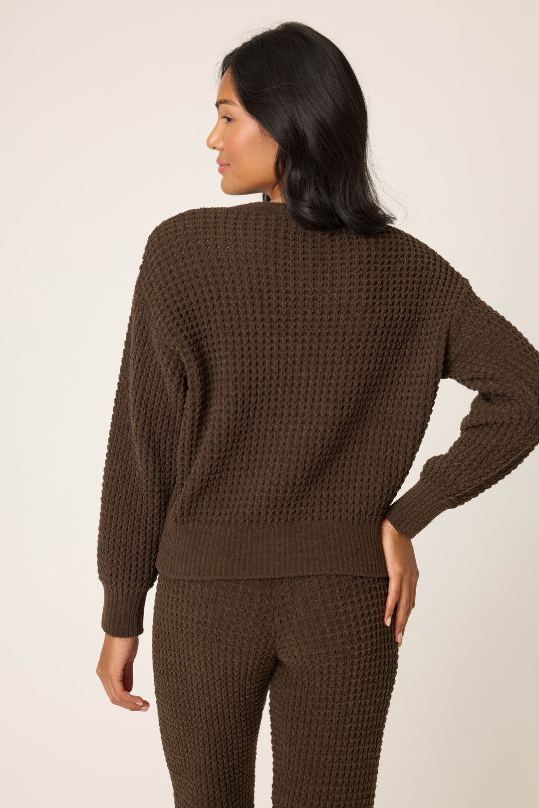 A woman faces away from the camera, wearing the P.J. Salvage Chenille Waffle Solid Chocolate Sweater with matching pants. Her right hand is on her hip, and she has medium-length dark hair. The background is plain and light-colored.