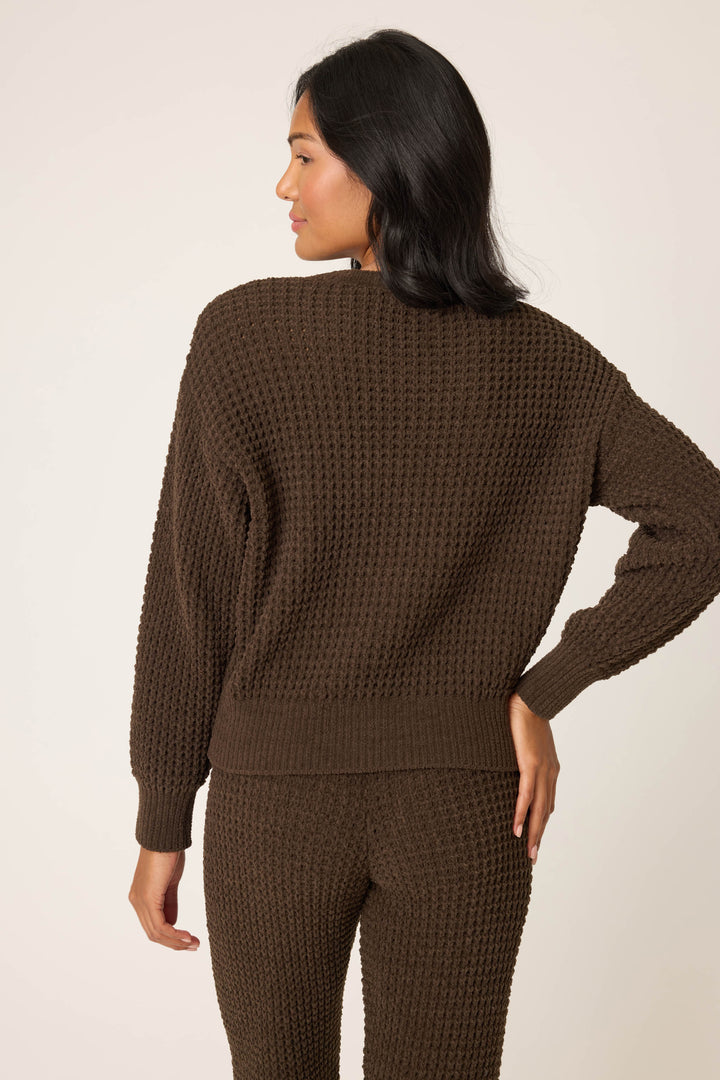 A woman faces away from the camera, wearing the P.J. Salvage Chenille Waffle Solid Chocolate Sweater with matching pants. Her right hand is on her hip, and she has medium-length dark hair. The background is plain and light-colored.