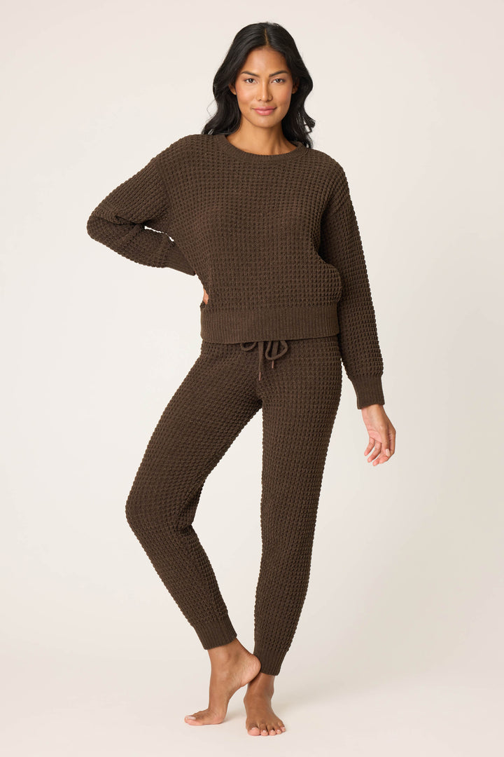 A person with long, dark hair wears the P.J. Salvage Chenille Waffle Solid Chocolate Sweater lounge set with a long-sleeve top and drawstring pants, standing barefoot against a plain light background.