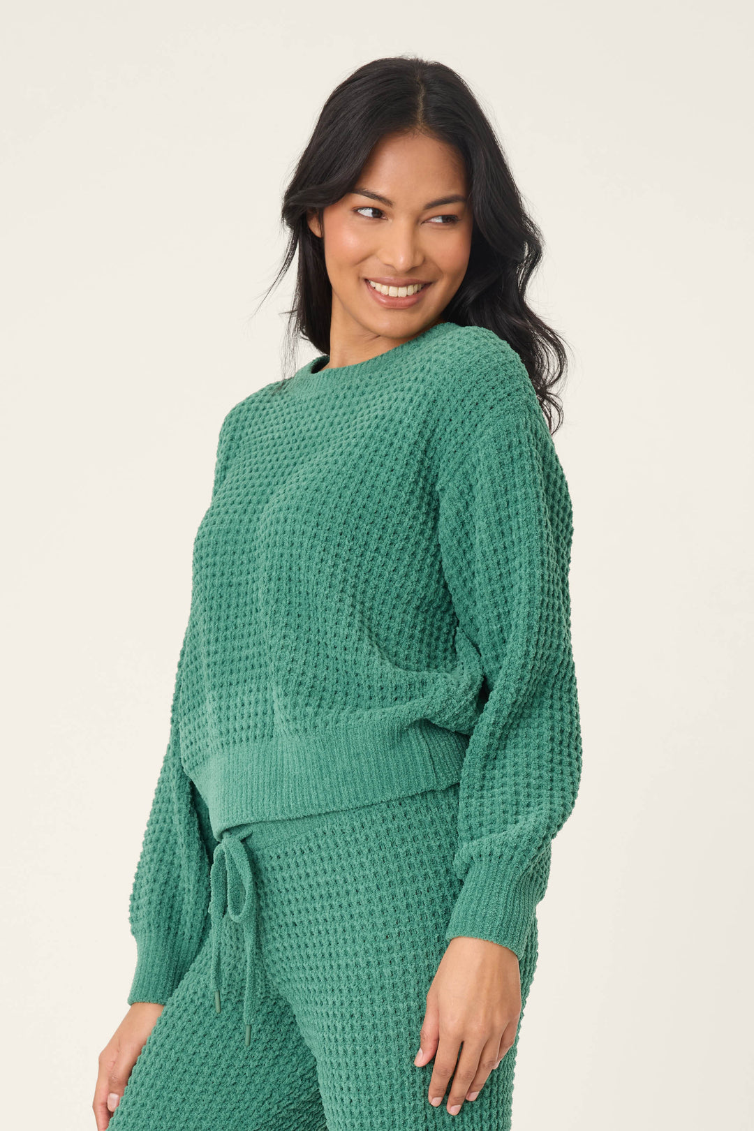 Wearing a P.J. Salvage Chenille Waffle Solid Forest Sweater and matching pants, a person stands against a light background, looking to the side and smiling.