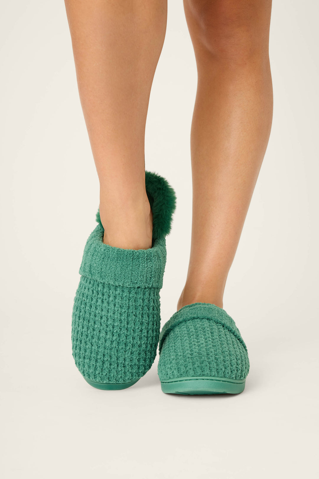 A person stands with feet together on a light background, wearing P.J. Salvage Chenille Waffle Cozy Slippers in teal green with textured fabric and a fluffy cuff. Only the legs and feet are shown.