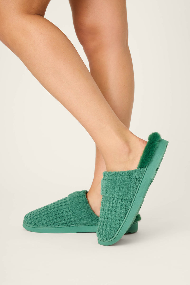 A person wears P.J. Salvage Chenille Waffle Cozy Slippers with a fuzzy lining, standing on a light-colored surface. Only their lower legs and feet are visible.
