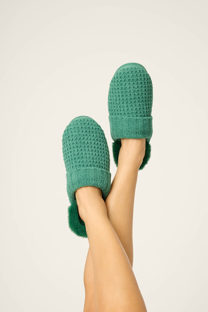 A person raises crossed legs in the air, showing P.J. Salvage Chenille Waffle Cozy Slippers—green knit with a textured pattern and fur trim—against a plain light background.