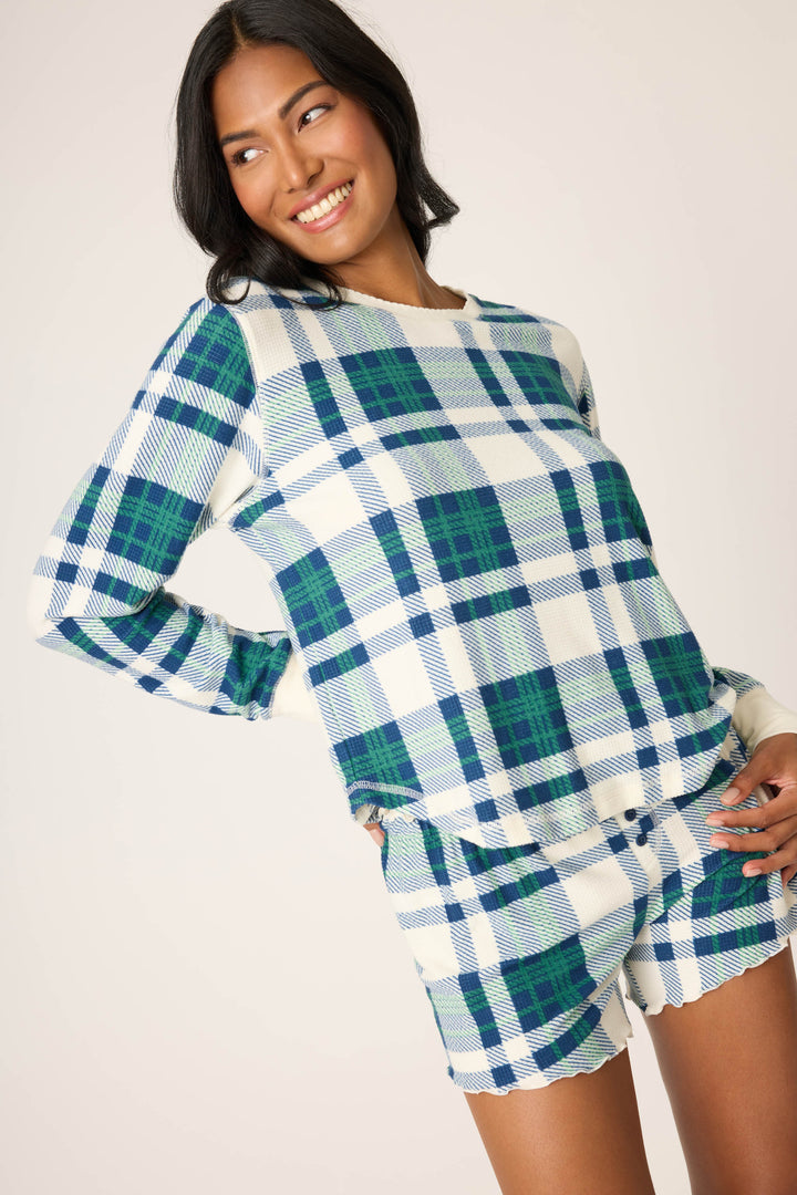 A woman smiles, wearing the P.J. Salvage Cozy Life Plaid Velour Thermal Long Sleeve Top in a blue, green, and white plaid pattern with matching shorts. The background is plain and light-colored.