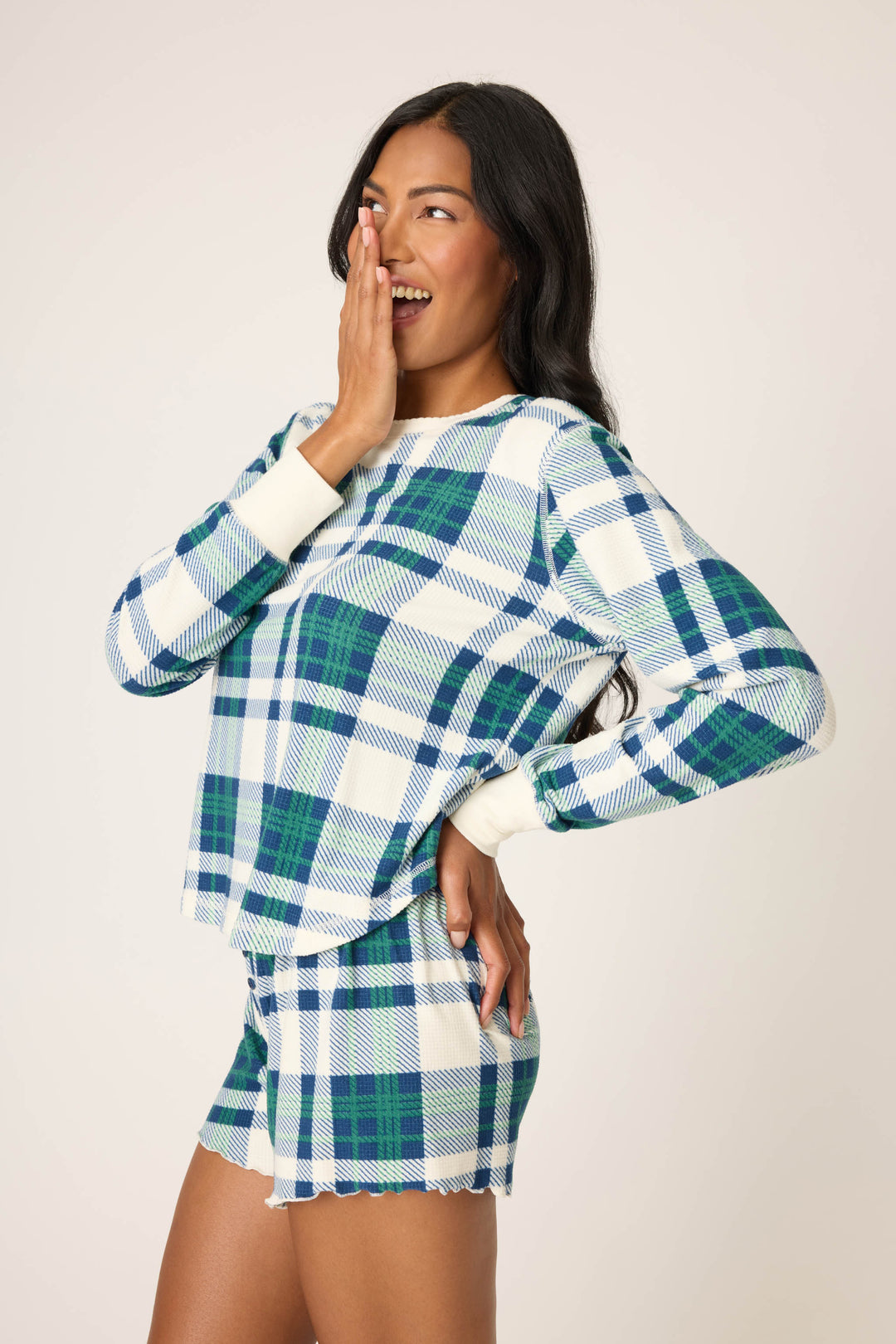 A woman wears the P.J. Salvage Cozy Life Plaid Velour Thermal Long Sleeve Top in blue, green, and white plaid, standing sideways with one hand on her hip and the other covering her smiling mouth as she looks to the side.