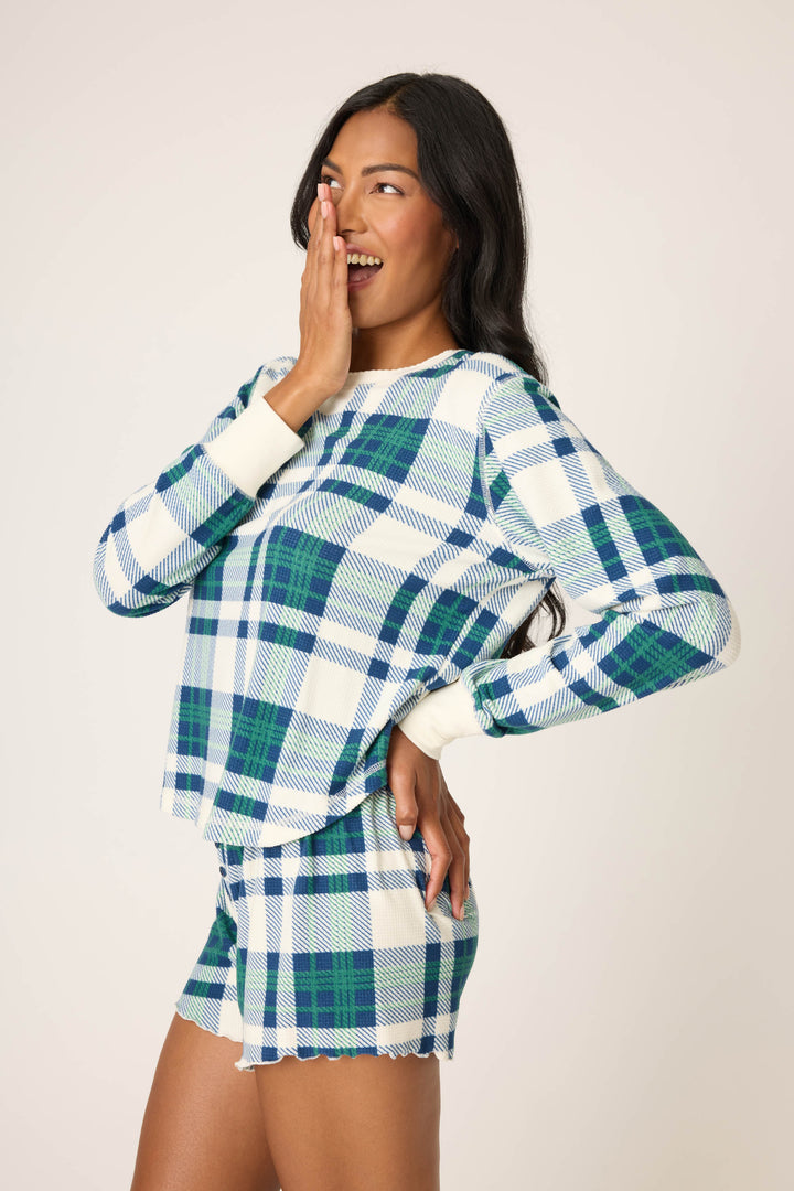 A woman wears the P.J. Salvage Cozy Life Plaid Velour Thermal Long Sleeve Top in blue, green, and white plaid, standing sideways with one hand on her hip and the other covering her smiling mouth as she looks to the side.