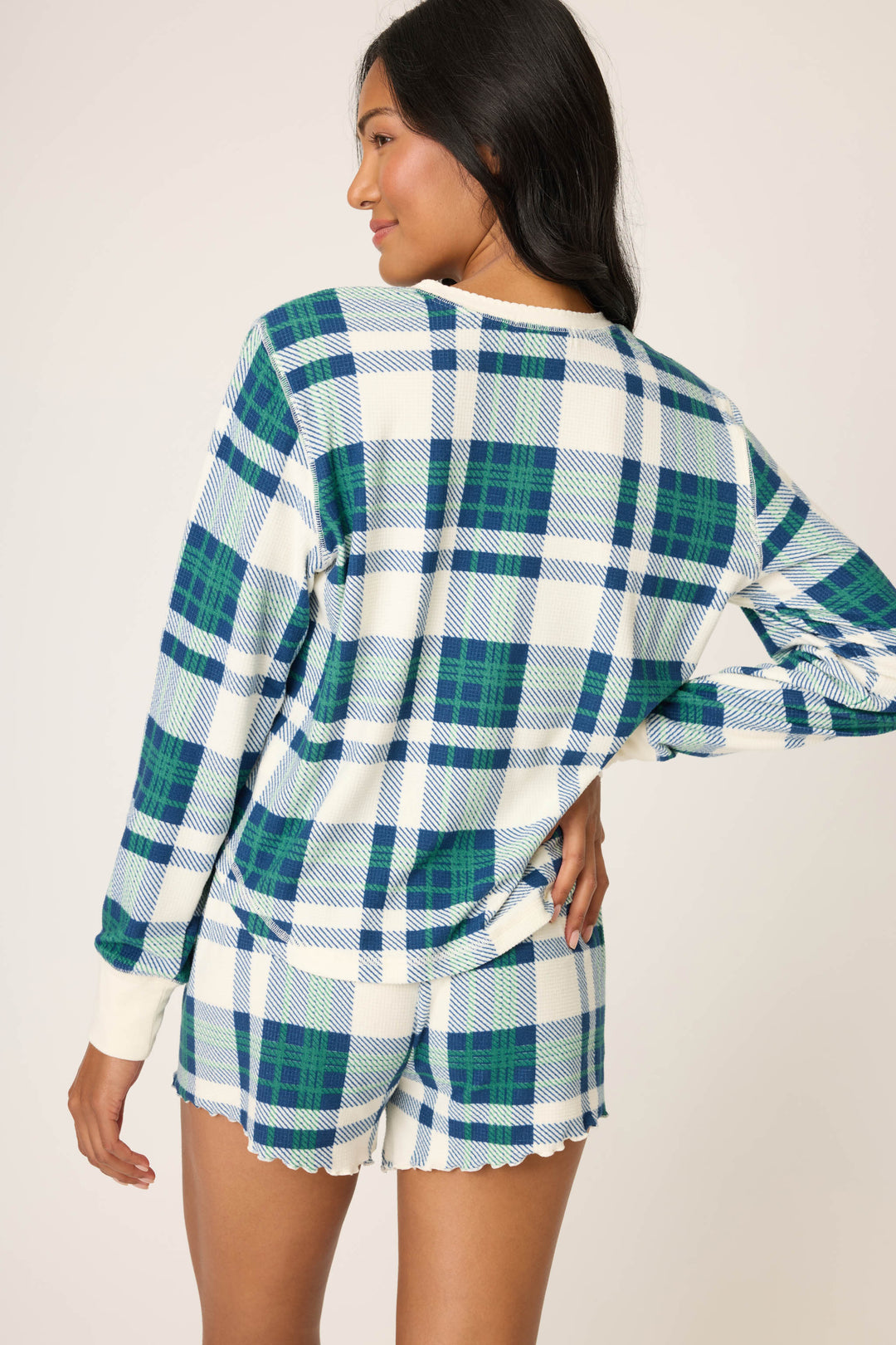 A person stands with their back to the camera, wearing the P.J. Salvage Cozy Life Plaid Velour Thermal Long Sleeve Top in a blue, green, and white plaid pattern. One hand is on their hip.