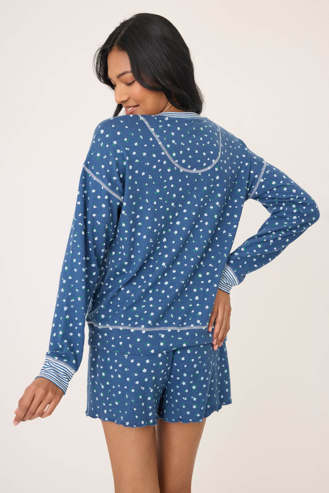 A woman with long dark hair faces away wearing the P.J. Salvage Cozy Life Floral Long Sleeve Top, a blue pajama with white and green floral patterns and light trim, paired with matching shorts. Her left hand is on her hip.
