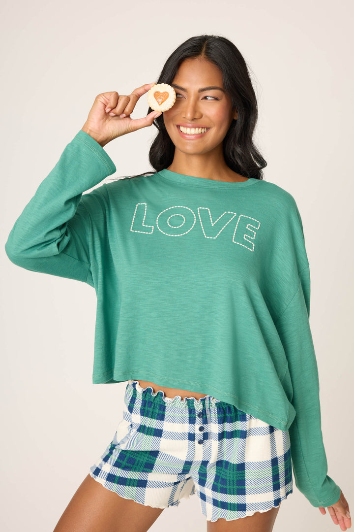 A woman with long dark hair smiles while holding a heart-shaped cookie over one eye. She wears the P.J. Salvage Cozy Life "Love" Graphic Long Sleeve Top and blue plaid shorts.