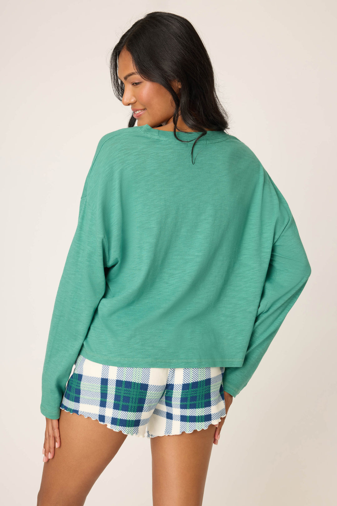 A person faces away, wearing the P.J. Salvage Cozy Life "Love" Graphic Long Sleeve Top in green, paired with blue-and-green plaid scalloped shorts, standing against a plain light background.