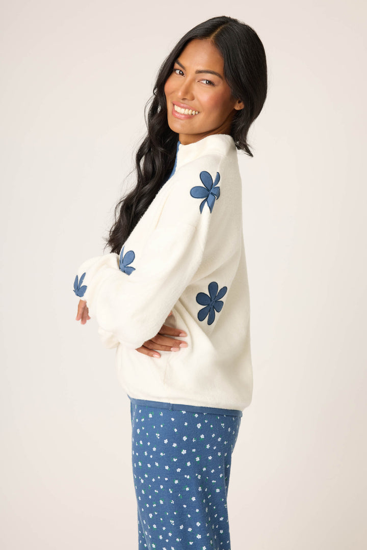 A woman with long dark hair smiles sideways in a light background, wearing the P.J. Salvage Cozy Life Floral Quarter-Zip Fleece Sweatshirt with blue floral sleeve designs and blue pants featuring a small white print.