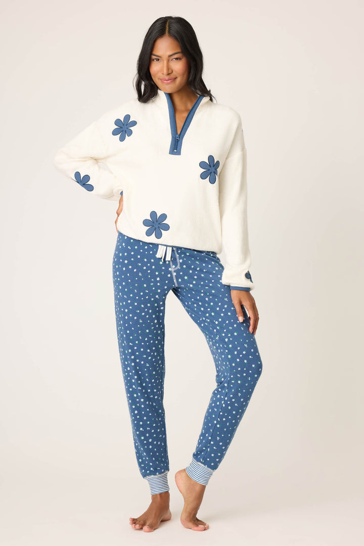 A person stands barefoot in blue polka dot jogger pants and the P.J. Salvage Cozy Life Floral Quarter-Zip Fleece Sweatshirt, featuring large blue flowers and a blue collar, posing against a plain background.