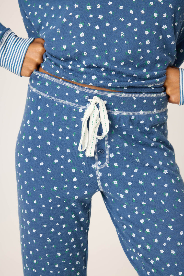 A person stands with hands on hips wearing the P.J. Salvage Cozy Life Floral Banded Pant, featuring a blue base with small white and green florals, striped cuffs, and a white drawstring waist.