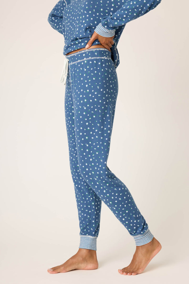 A person stands barefoot, wearing the P.J. Salvage Cozy Life Floral Banded Pant. Their hand rests on their hip, with a glimpse of the matching pajama top visible. The background is plain and light-colored.