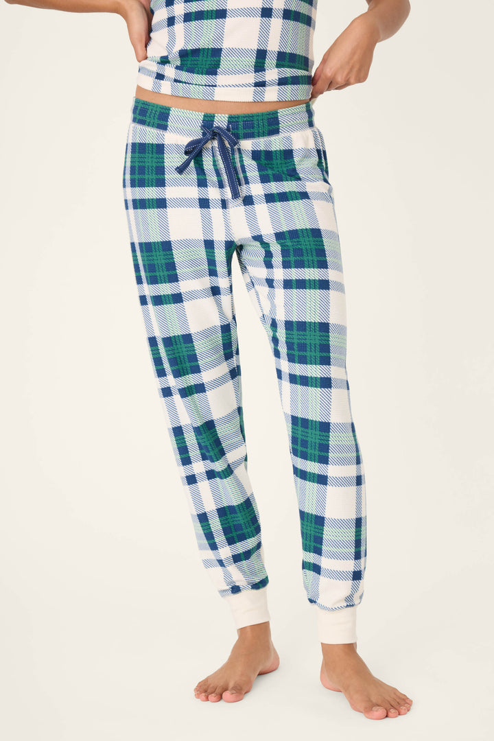 A person models the P.J. Salvage Cozy Life Plaid Velour Thermal Banded Pant, featuring blue, green, and white plaid with a drawstring waist and pockets, standing barefoot against a plain light background.