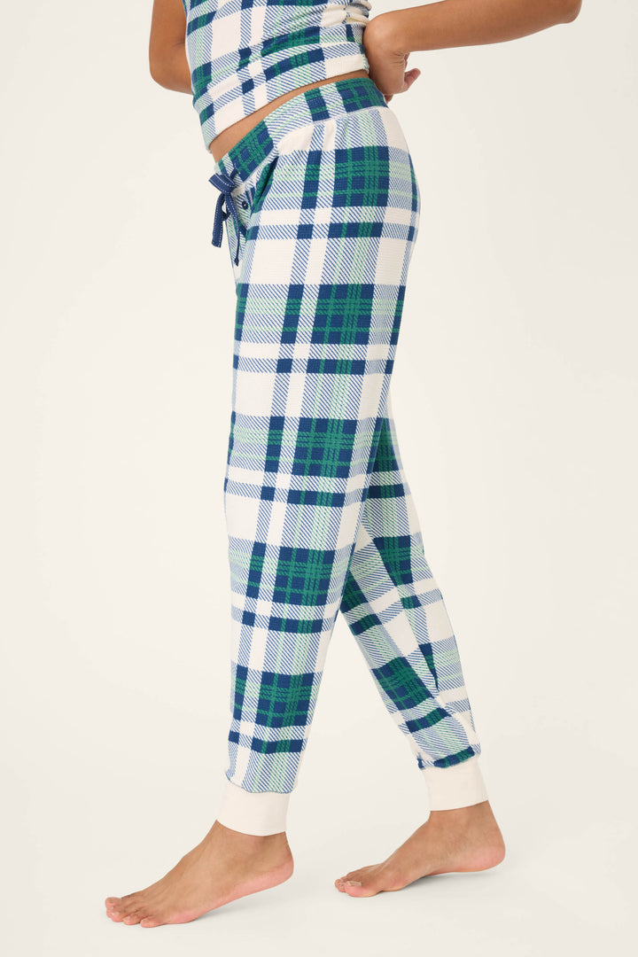 A person stands barefoot in P.J. Salvage's Cozy Life Plaid Velour Thermal Banded Pant, featuring a blue, green, and white plaid pattern with a drawstring waist and white cuffs, shown in a side view against a plain background.