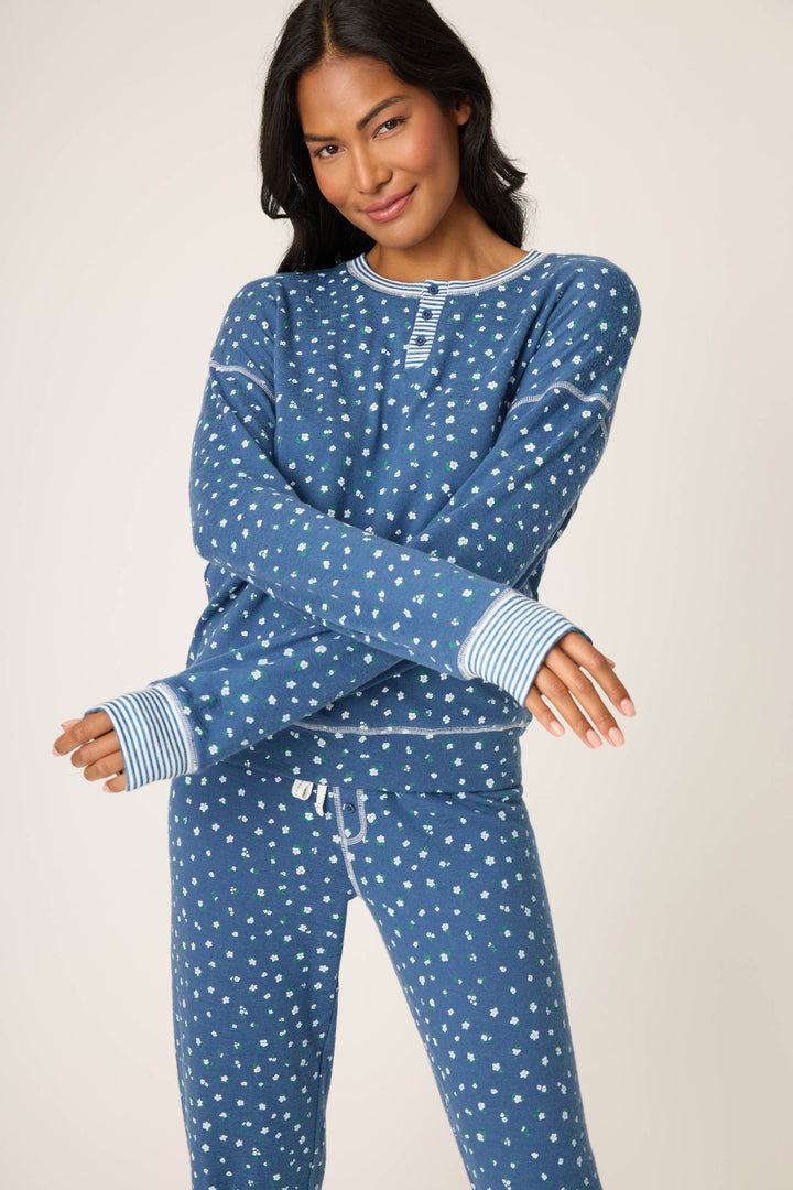 A woman poses with arms crossed, wearing the Cozy Life Floral PJ Set by P.J. Salvage—a matching blue pajama set with a small white floral design and striped cuffs—against a plain light background.