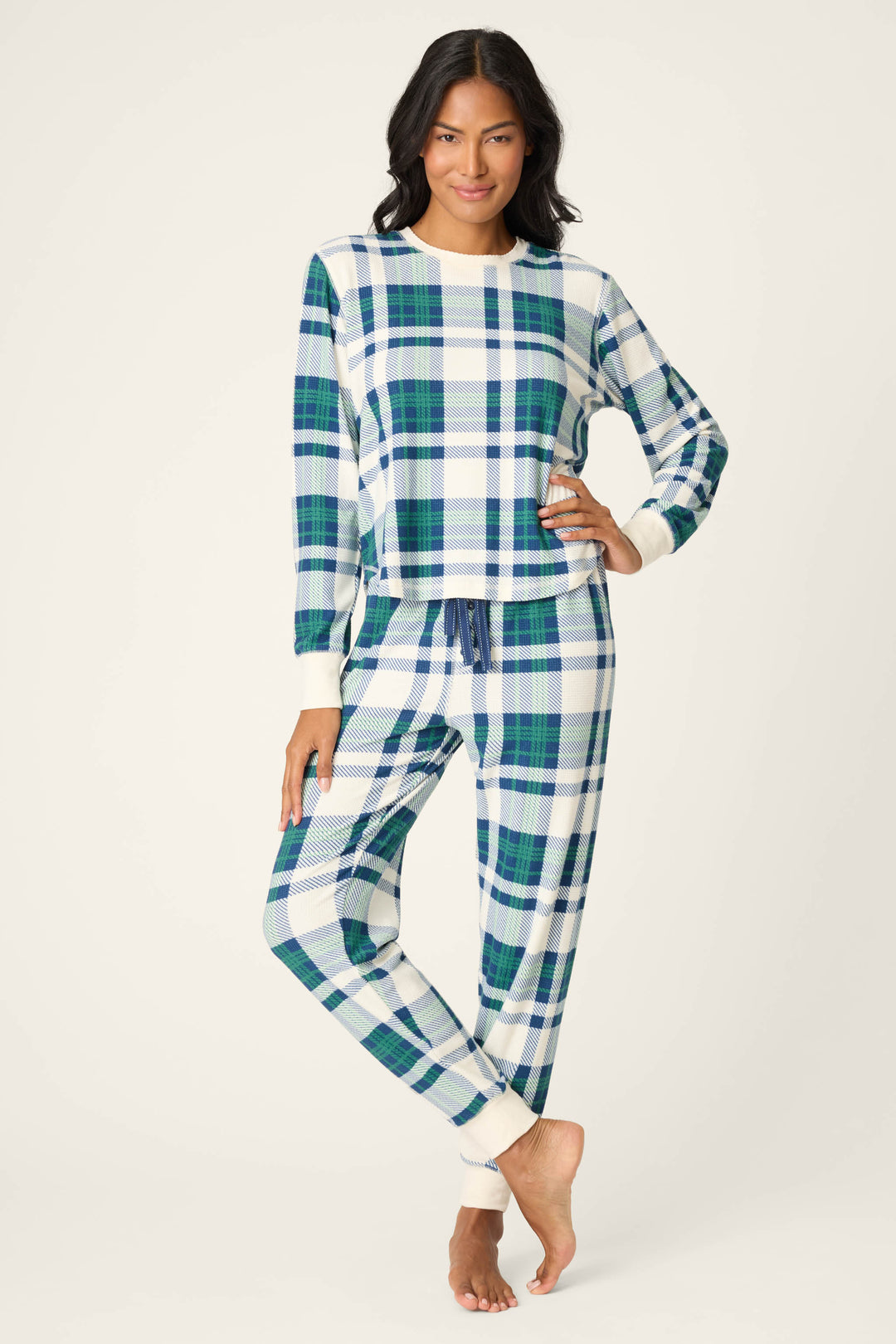 A person stands barefoot against a plain background, wearing the P.J. Salvage Cozy Life Plaid Velour Thermal PJ Set—long-sleeved with a blue, green, and white plaid print and drawstring waist. One hand is on their hip, the other by their side.