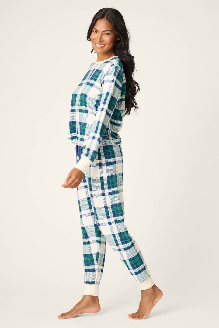 A woman stands barefoot in profile wearing the P.J. Salvage Cozy Life Plaid Velour Thermal PJ Set—blue and green plaid with white cuffs—posing against a plain light background.