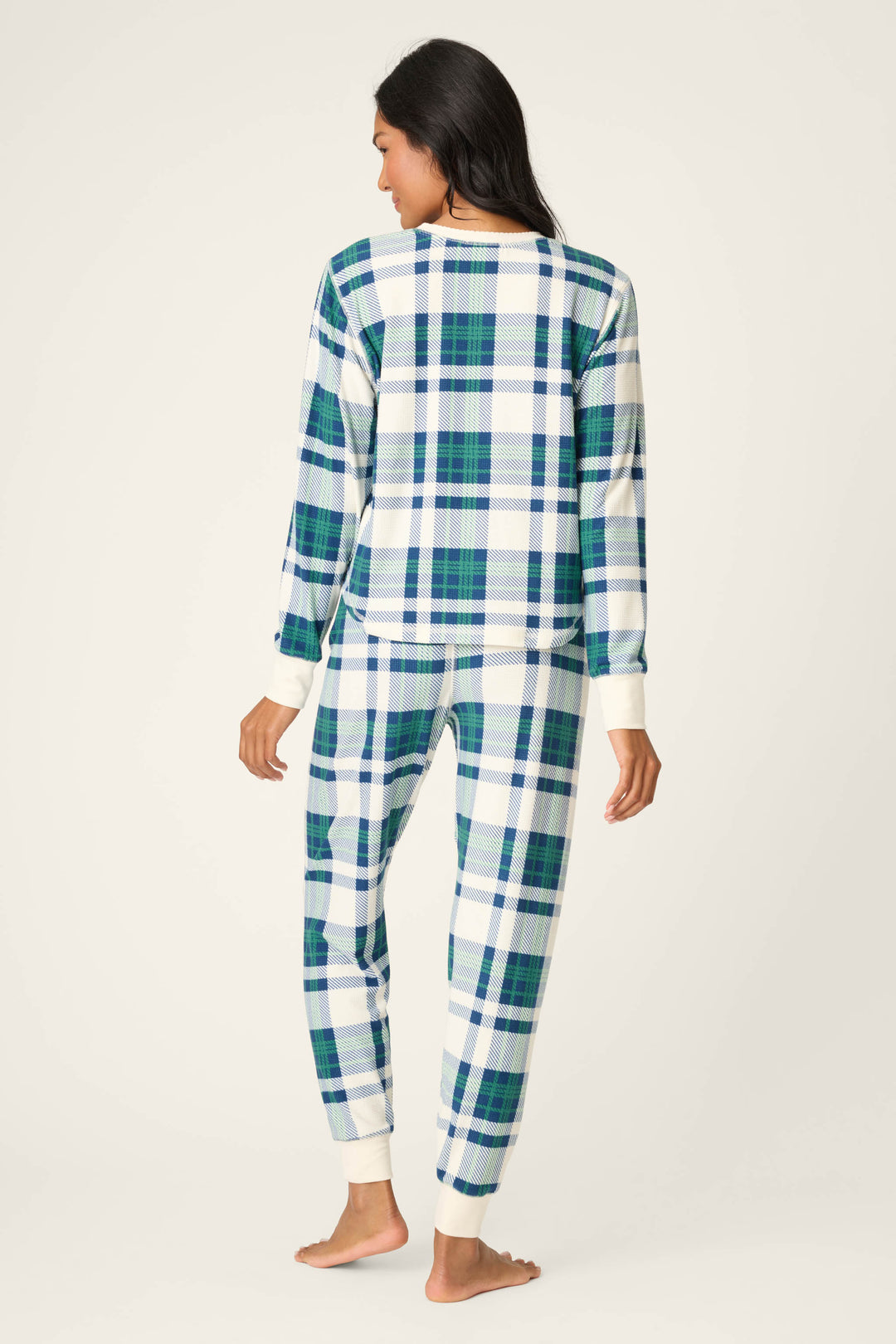A person stands barefoot and facing away in a P.J. Salvage Cozy Life Plaid Velour Thermal PJ Set, featuring blue, green, and white plaid with cream cuffs on the sleeves and ankles. The background is plain and light-colored.