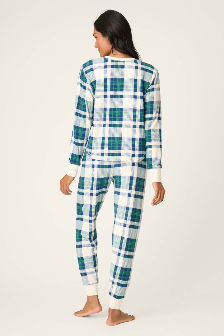 A person stands barefoot and facing away in a P.J. Salvage Cozy Life Plaid Velour Thermal PJ Set, featuring blue, green, and white plaid with cream cuffs on the sleeves and ankles. The background is plain and light-colored.