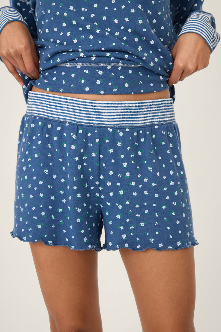 A person wears P.J. Salvage Cozy Life Floral Shorts—blue with a small white and green floral print and a striped elastic waistband. The matching top is partly visible as they hold the shirt near their waist.