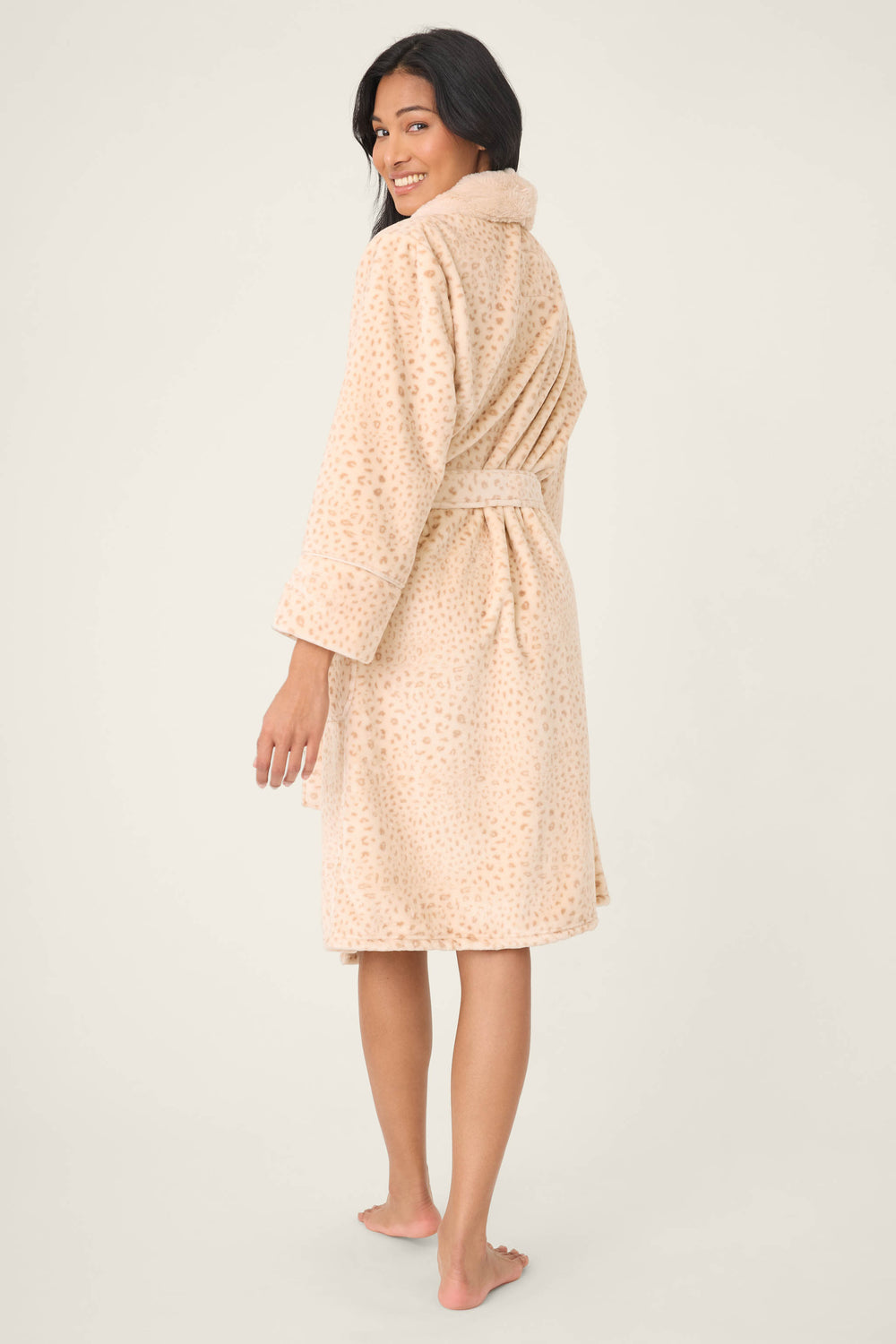 A barefoot woman faces backwards, smiling at the camera, wearing the P.J. Salvage Luxe Plush Leopard Print Robe—a beige, knee-length robe with long sleeves, a tie belt, and a subtle animal print pattern.
