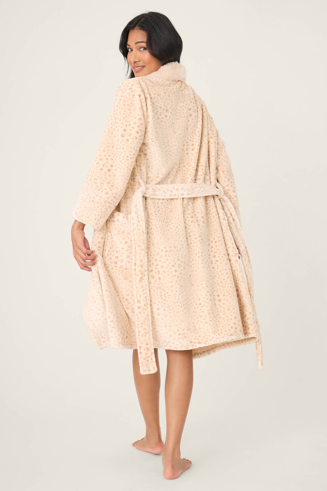 A woman stands barefoot, looking over her shoulder in the knee-length P.J. Salvage Luxe Plush Leopard Print Robe, featuring a soft beige fabric with a subtle leopard pattern and waist tie. The background is plain and light-colored.