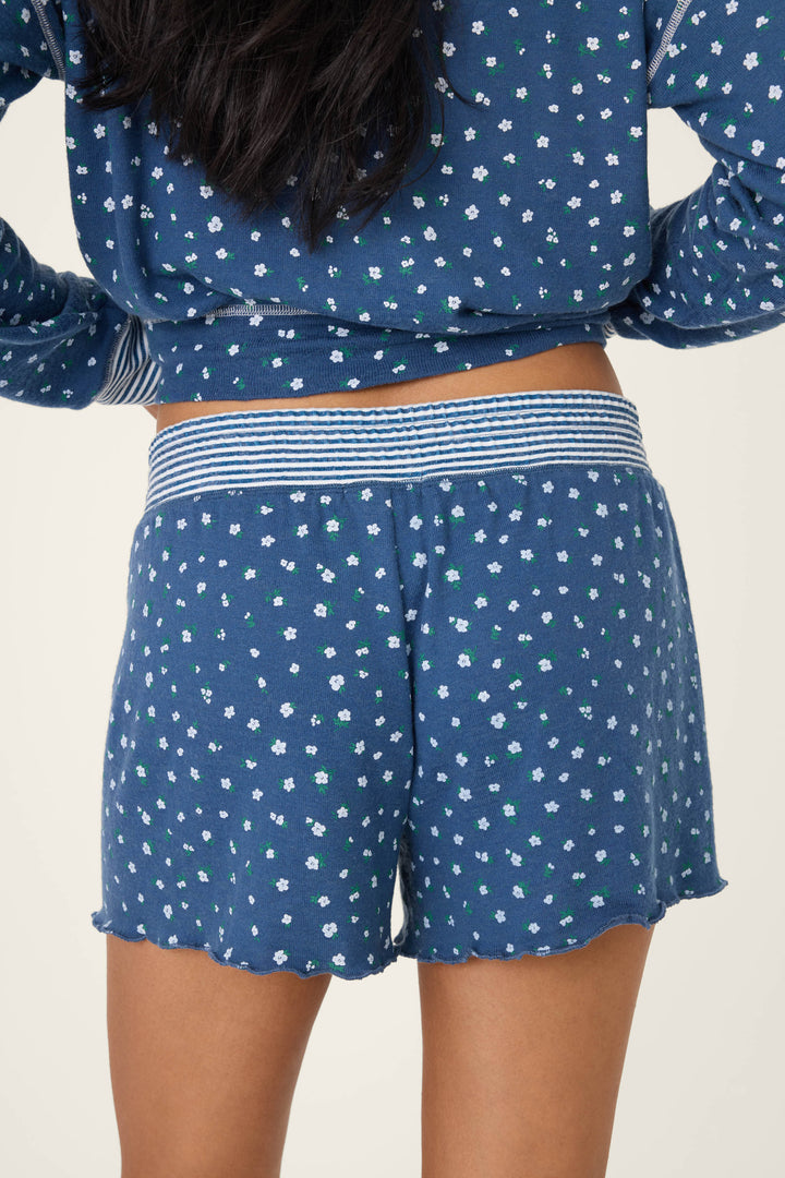 A person stands with their back to the camera, wearing P.J. Salvage’s Cozy Life Floral Short—blue pajama shorts featuring a small floral print and a striped waistband. The matching top is partially visible.