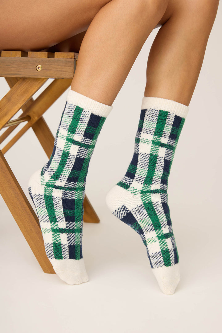 A person sits on a wooden chair with legs crossed, wearing P.J. Salvage Cozy Life Plaid Crew Socks in white with green, blue, and navy plaid. The background is plain and light-colored.