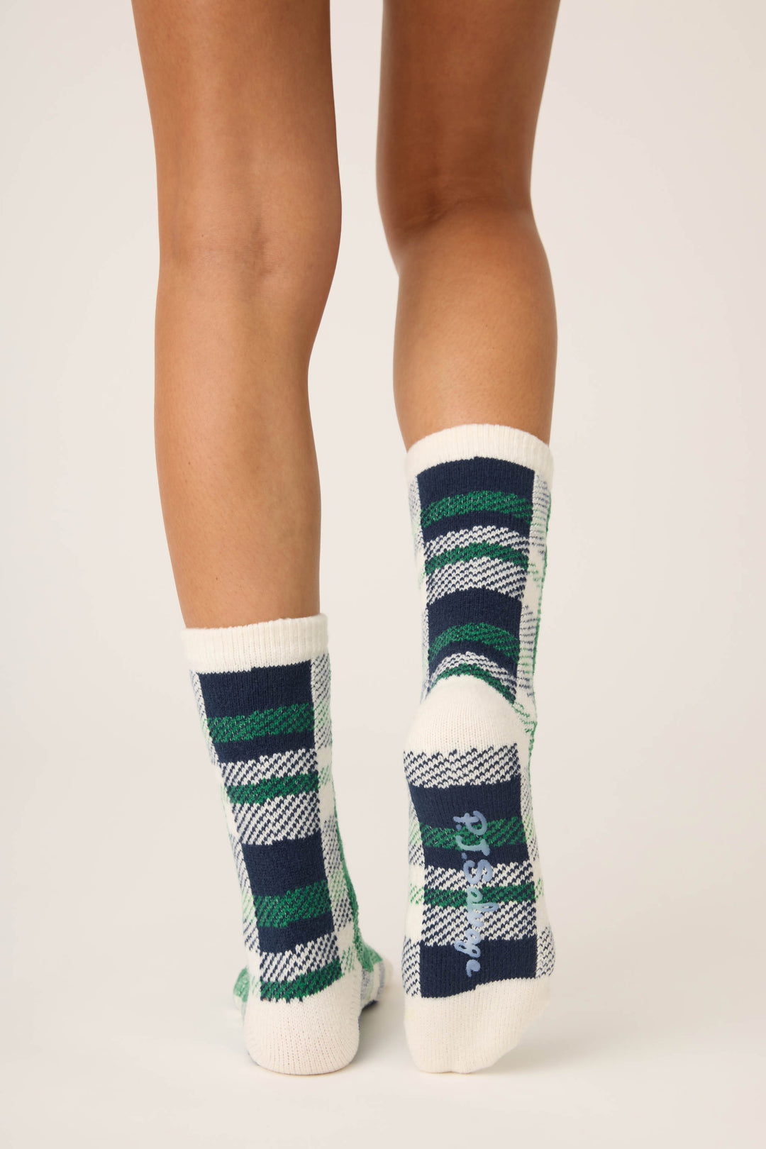 A person stands with their back to the camera, wearing P.J. Salvage Cozy Life Plaid Crew Socks in green, navy, and white plaid. "Dreamer" is written in cursive on the sole of the right sock.