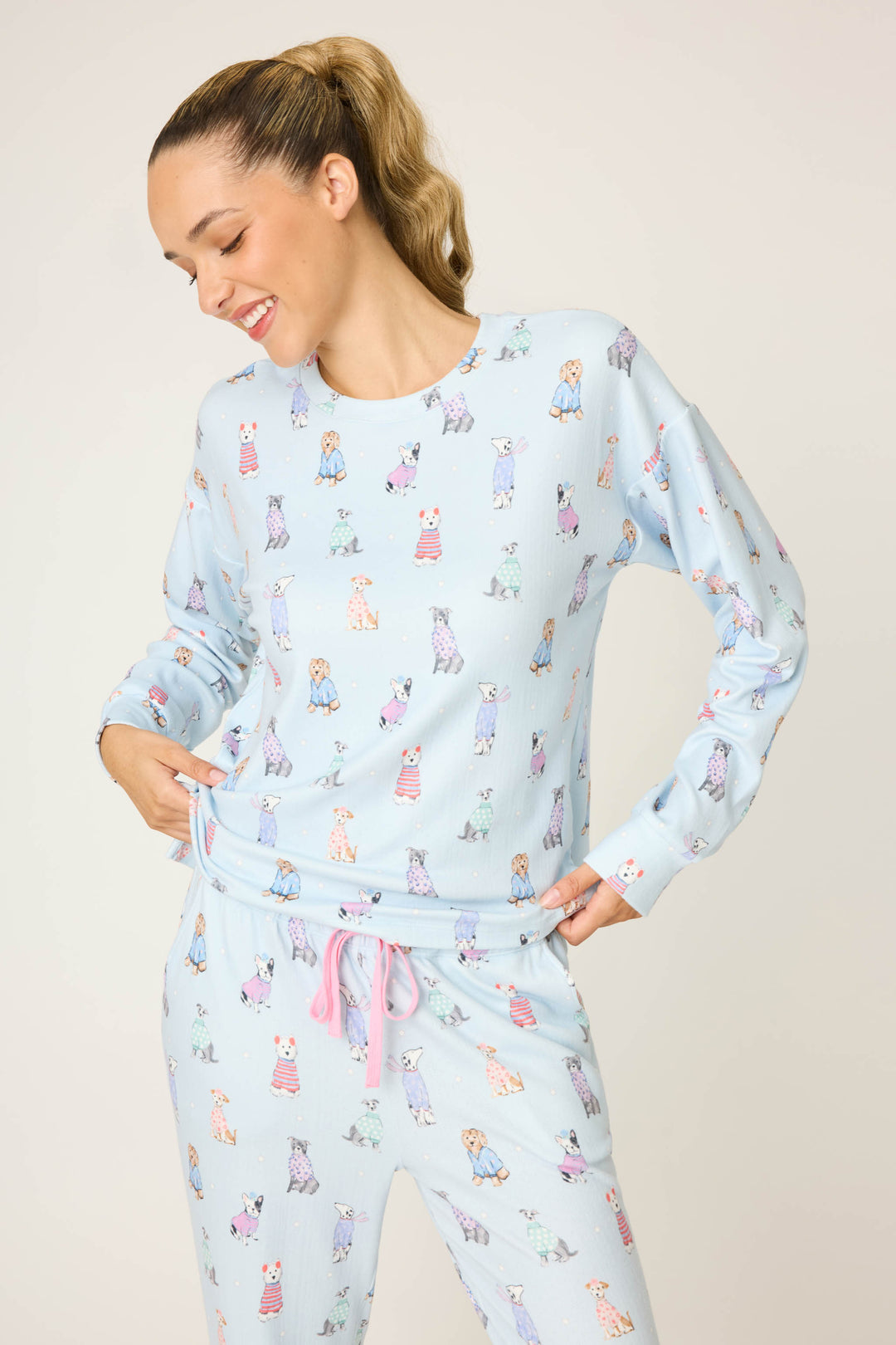 A person stands against a plain light background, smiling and looking down with one hand on their waist. They are wearing the P.J. Salvage Doggone Cold Dog Print Pointelle Long Sleeve Top in light blue with cartoon dog prints and a pink drawstring.