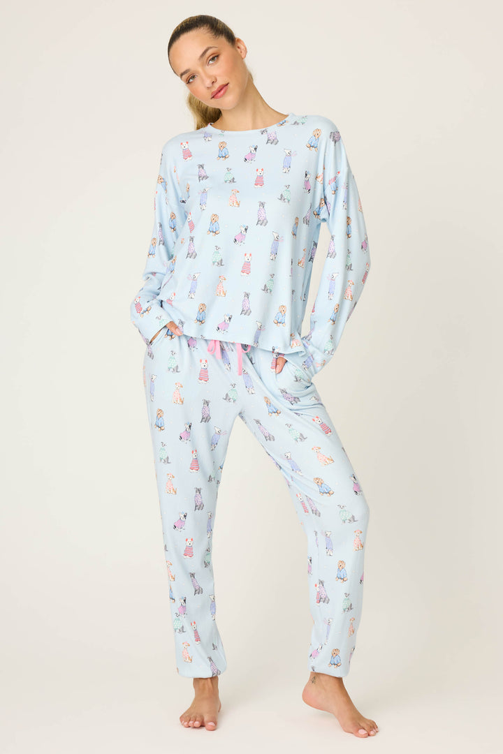 A person stands barefoot against a light background, wearing the P.J. Salvage Doggone Cold Dog Print Pointelle Long Sleeve Top and relaxed-fit pants in light blue with colorful cartoon dog prints.