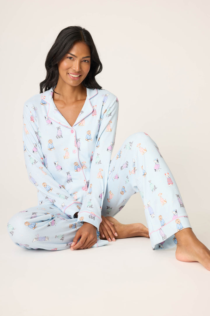 A person with long dark hair wears the P.J. Salvage Doggone Cold Dog Print Pointelle PJ Set, sitting cross-legged on the floor and smiling at the camera against a plain light background.