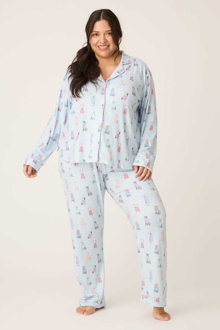A woman smiles at the camera, barefoot in the P.J. Salvage Doggone Cold Dog Print Pointelle Plus Size PJ Set—light blue with a colorful dog print, featuring pajama pants and a matching long-sleeve button-up top.