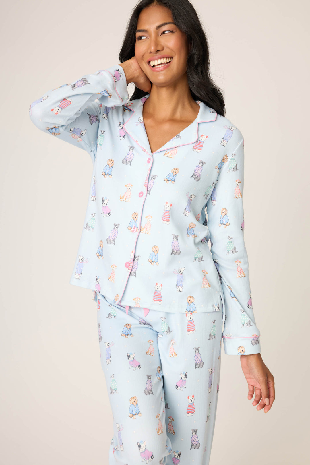 A person smiles and touches their face while wearing the P.J. Salvage Doggone Cold Dog Print Pointelle PJ Set, featuring light blue long-sleeve button-up top and matching pants adorned with colorful cartoon dogs.