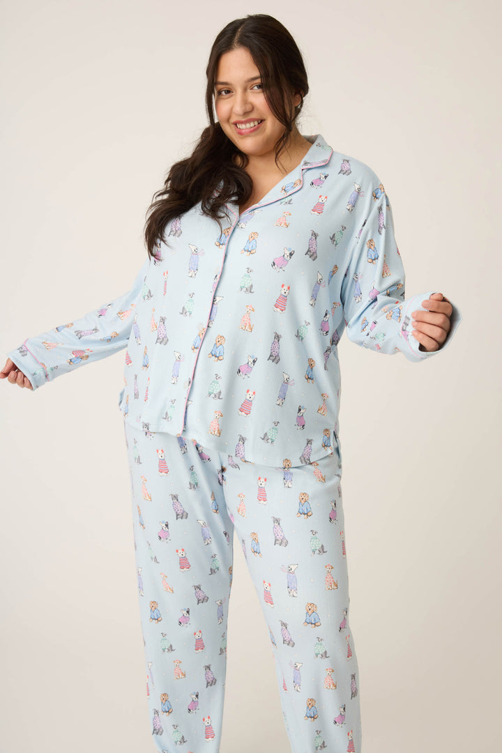 A woman with long dark hair wears the P.J. Salvage Doggone Cold Dog Print Pointelle Plus Size PJ Set, featuring light blue pajamas with colorful dog prints, standing against a plain light background and smiling with her arms slightly raised.
