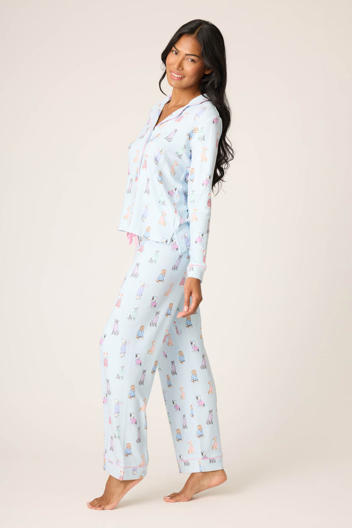 A woman stands barefoot against a plain background, smiling sideways while modeling the Doggone Cold Dog Print Pointelle PJ Set by P.J. Salvage, featuring light blue pajamas decorated with colorful cartoon dog prints.