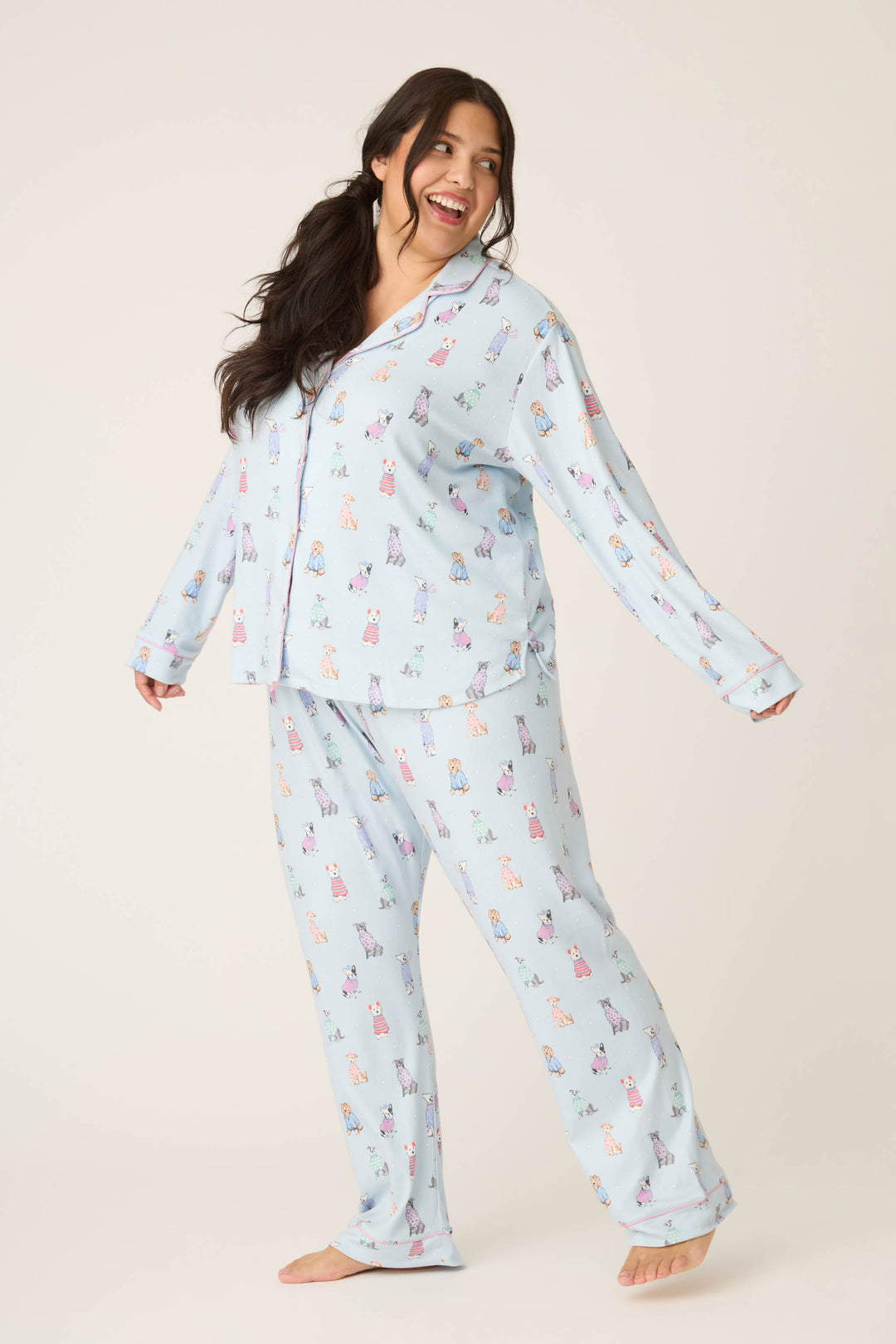 A woman with long dark hair smiles and looks to the side, barefoot on a plain background, wearing the P.J. Salvage Doggone Cold Dog Print Pointelle Plus Size PJ Set in light blue with a colorful dog print pattern.