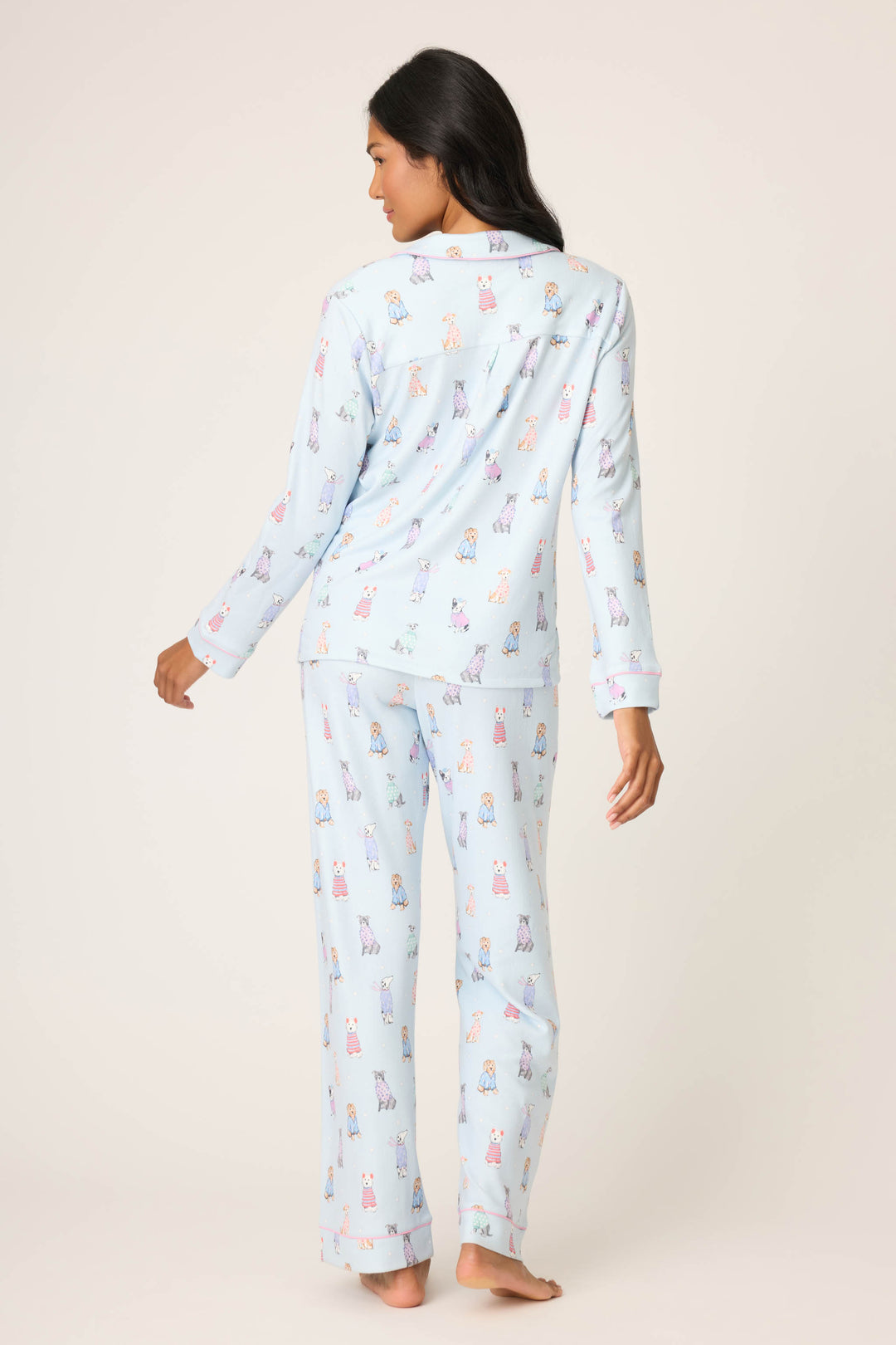A woman stands barefoot on a neutral background, facing away, wearing the P.J. Salvage Doggone Cold Dog Print Pointelle PJ Set—a light blue, long-sleeved pajama set with colorful dog prints. Her long dark hair flows down her back.
