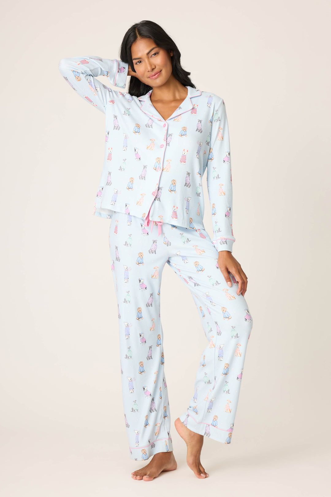 A woman stands barefoot against a light background, smiling and wearing the P.J. Salvage Doggone Cold Dog Print Pointelle PJ Set with pink piping, her hand resting on her head.
