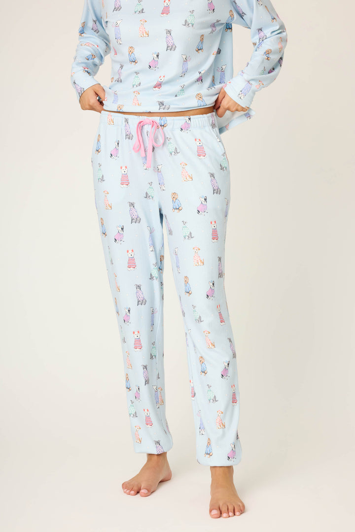 A person stands barefoot against a plain background, wearing P.J. Salvage Doggone Cold Dog Print Pointelle Banded Pants in light blue with a pink drawstring and a matching long-sleeve top featuring playful dog prints.