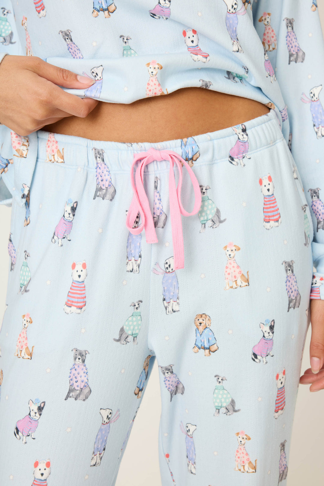 A person wears P.J. Salvage Doggone Cold Dog Print Pointelle Banded Pants in light blue, featuring dogs in various outfits, and lifts their pajama top slightly to reveal part of their midriff.