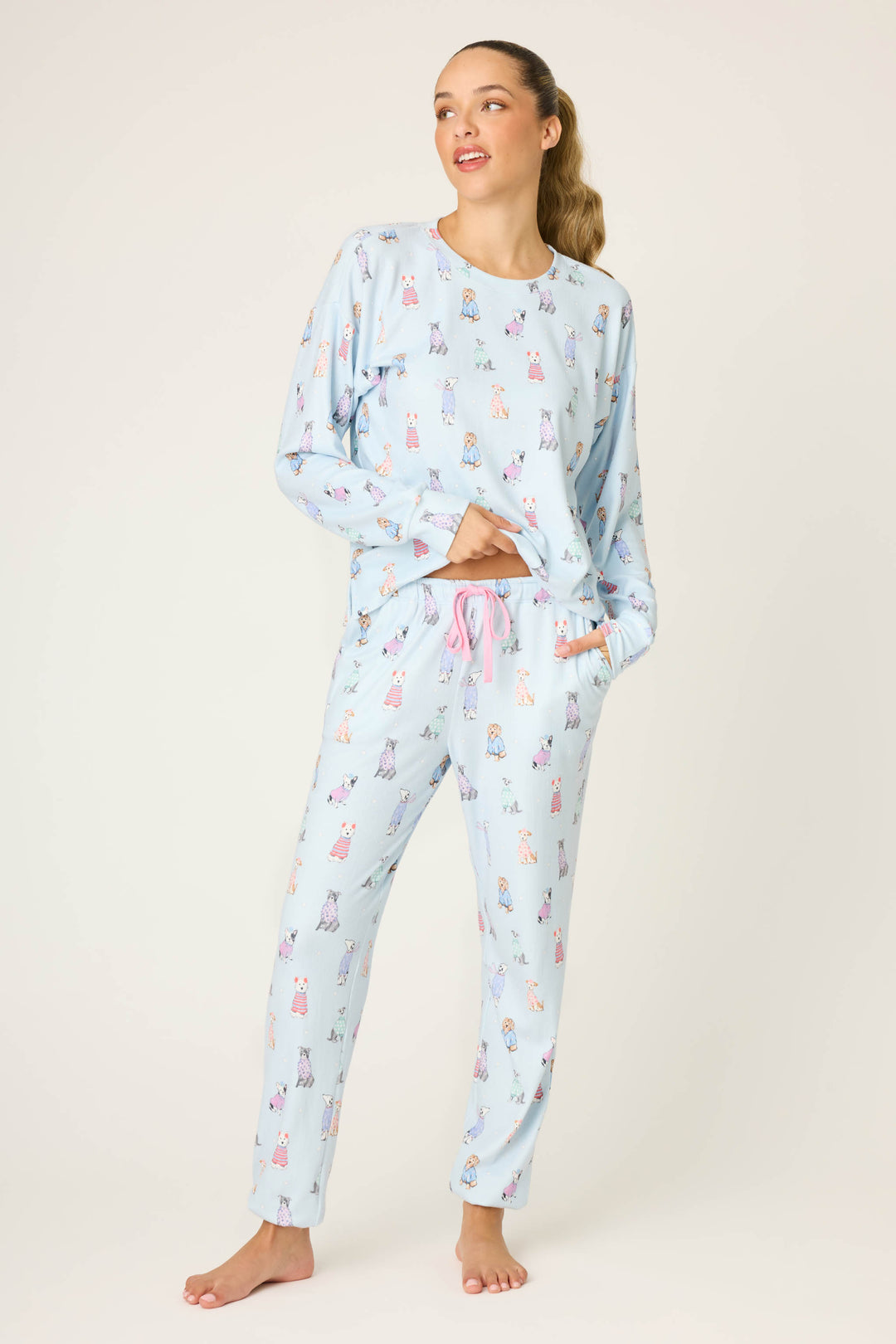 A woman stands barefoot against a light background, smiling as she looks to the side. She wears P.J. Salvage Doggone Cold Dog Print Pointelle Banded Pants—ideal winter loungewear—with one hand in her pocket.