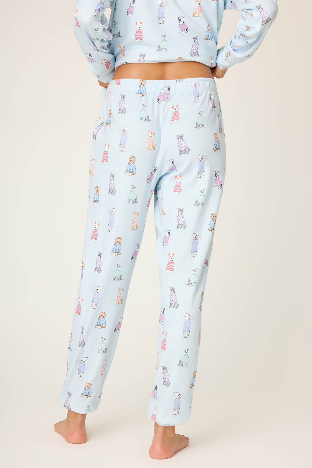 A person stands barefoot, facing away, wearing P.J. Salvage Doggone Cold Dog Print Pointelle Banded Pants—light blue with playful dog prints—paired with a matching top for cozy winter loungewear.