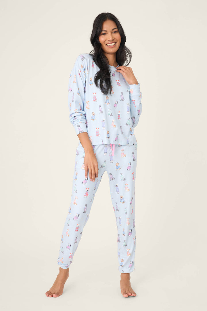 A person stands barefoot against a plain background, smiling with one hand near their neck. They wear the P.J. Salvage Doggone Cold Dog Print Pointelle Lounge Set—light blue, long-sleeve top and full-length pants with a colorful dog print.