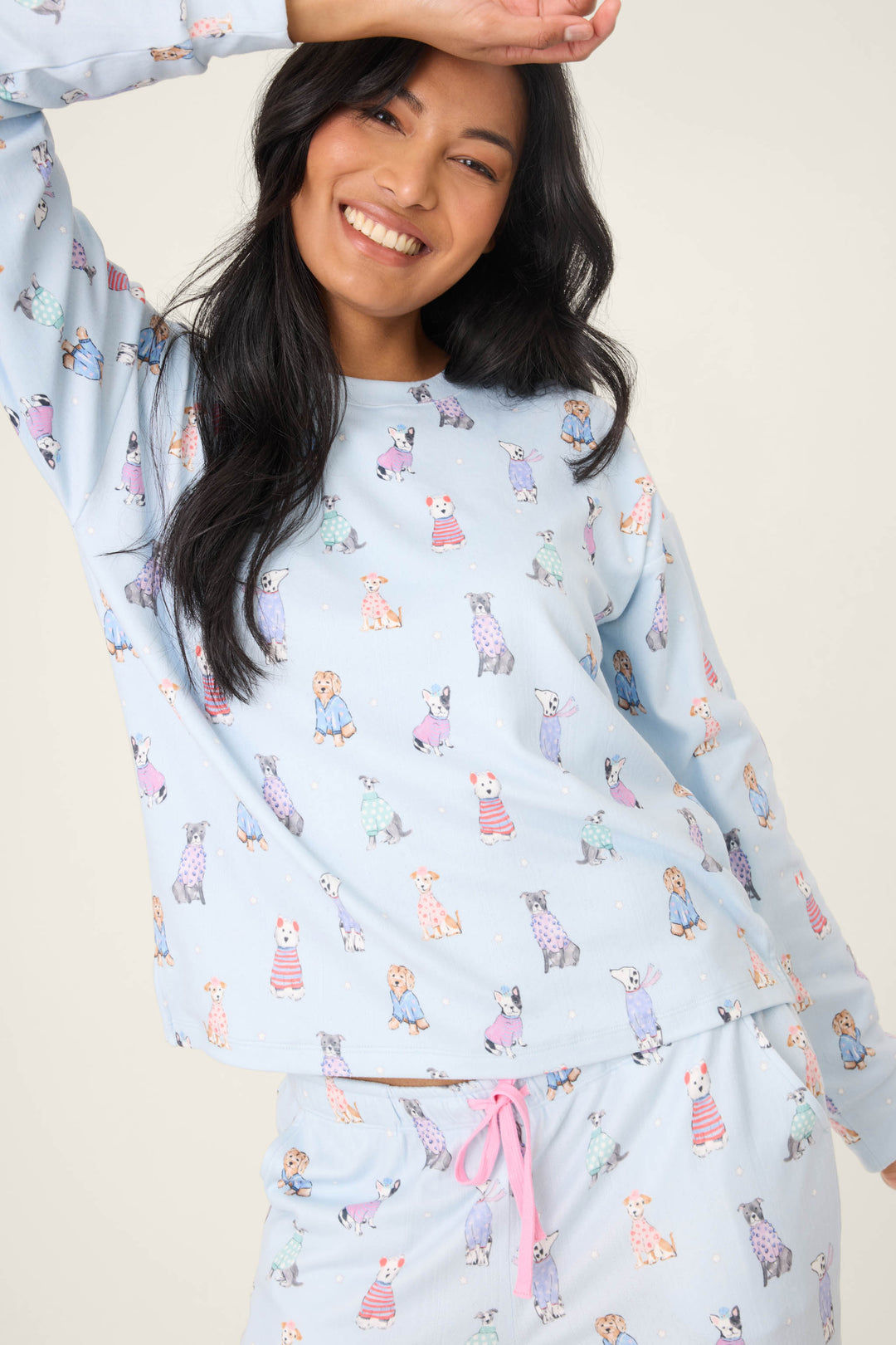 A woman with long black hair smiles and raises her left arm, wearing the P.J. Salvage Doggone Cold Dog Print Pointelle Lounge Set—light blue pajamas with a colorful dog print and pink drawstring—against a plain light background.