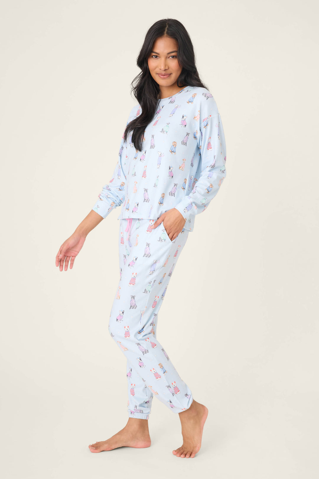 A person stands barefoot on a light background wearing the P.J. Salvage Doggone Cold Dog Print Pointelle Lounge Set, featuring playful dog prints in light blue with long sleeves and pants. One hand is in their pocket as they smile slightly.