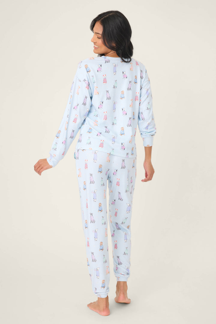 A woman stands barefoot on a light background, facing away and looking over her shoulder. She wears the P.J. Salvage Doggone Cold Dog Print Pointelle Lounge Set featuring a colorful cartoon dog pattern.